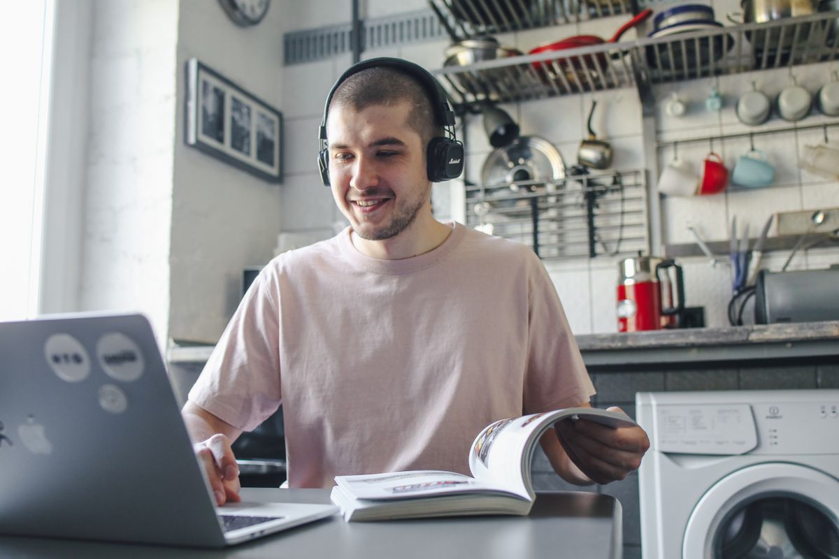 A young man wearing headphones studies online on a laptop at his kitchen table at home
