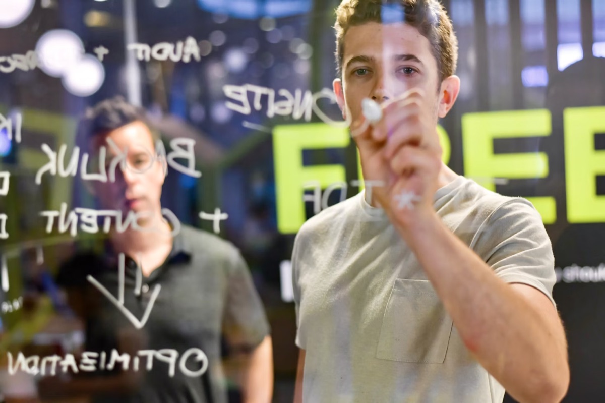 A student writes marketing strategy notes on a glass board while a colleague looks on