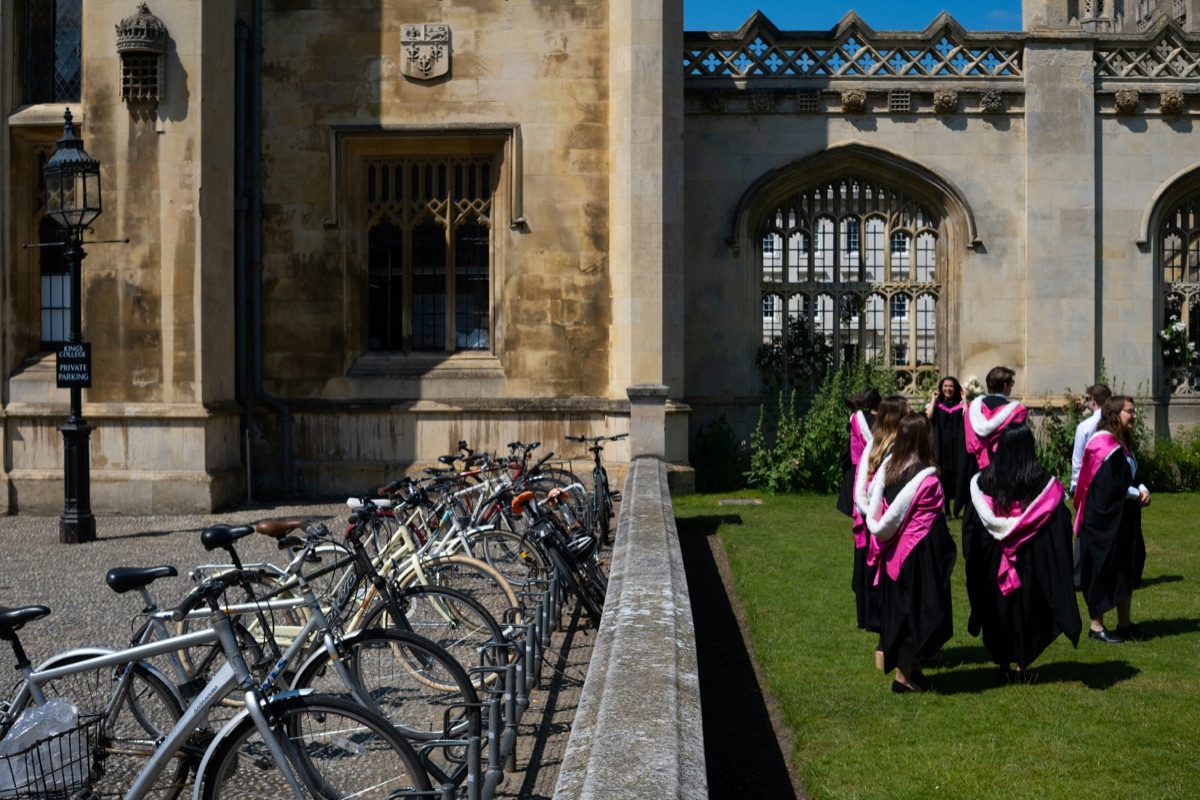 Graduates in gowns outside a historic college building in the UK, with bicycles parked along a wall